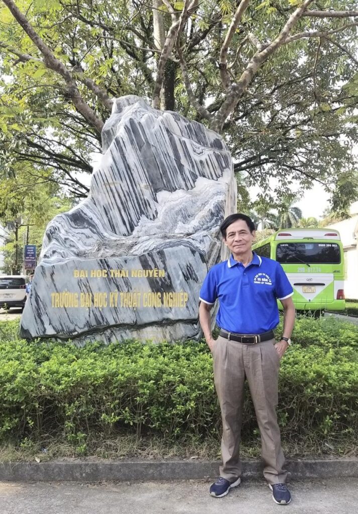 Nhà văn Nguyễn Trọng Luân đứng trước cổng đá Trường Đại học Kỹ thuật Công nghiệp - Đại học Thái Nguyên.